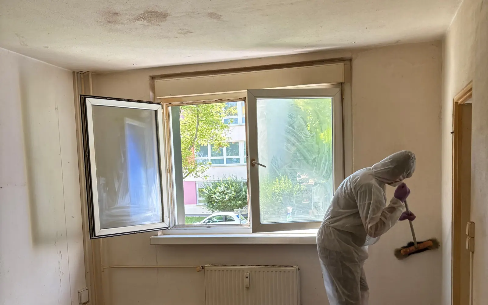 Mitarbeiter im weißen Schutzanzug fegt bei geöffnetem Fenster ein leer geräumtes Zimmer einer Berliner Wohnung besenrein