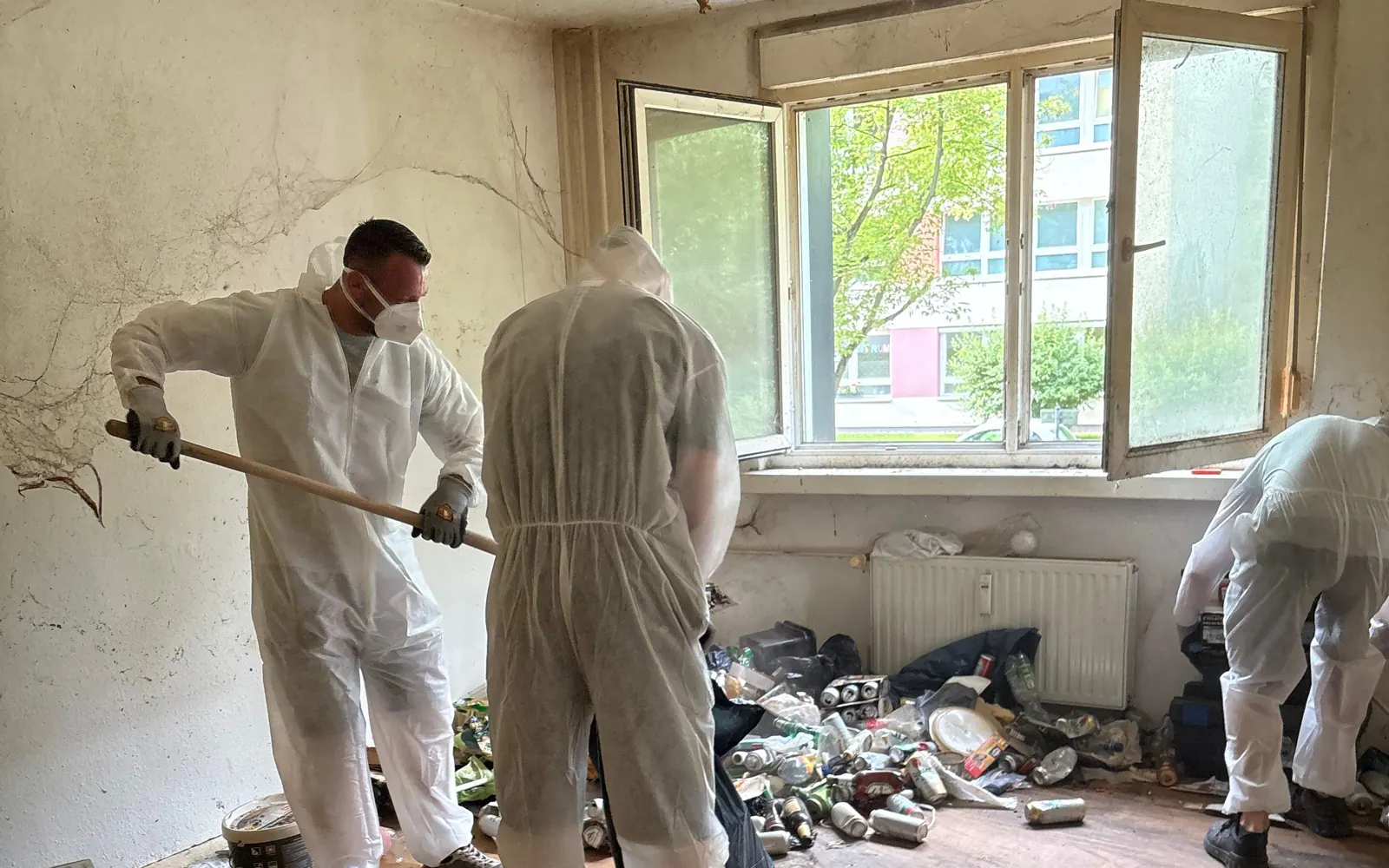 Zwei Mitarbeiter im weißen Schutzanzug räumen bei geöffnetem Fenster eine vermüllte Messie-Wohnung in Berlin