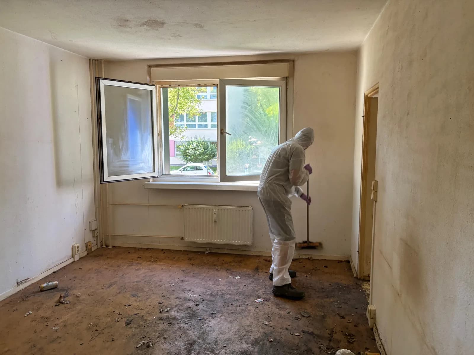 Mitarbeiter im weißen Schutzanzug fegt ein verschmutztes, leer geräumtes Zimmer bei einer Messie-Entrümpelung in Berlin
